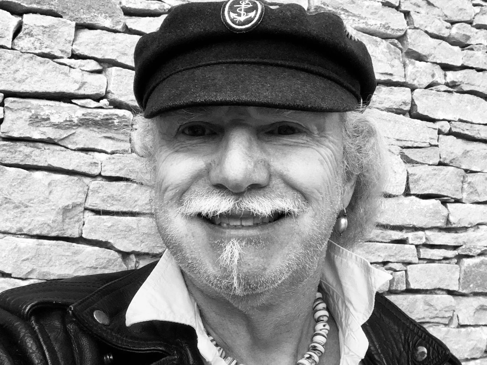 Un homme souriant, coiffé d'une casquette, devant un mur de pierre